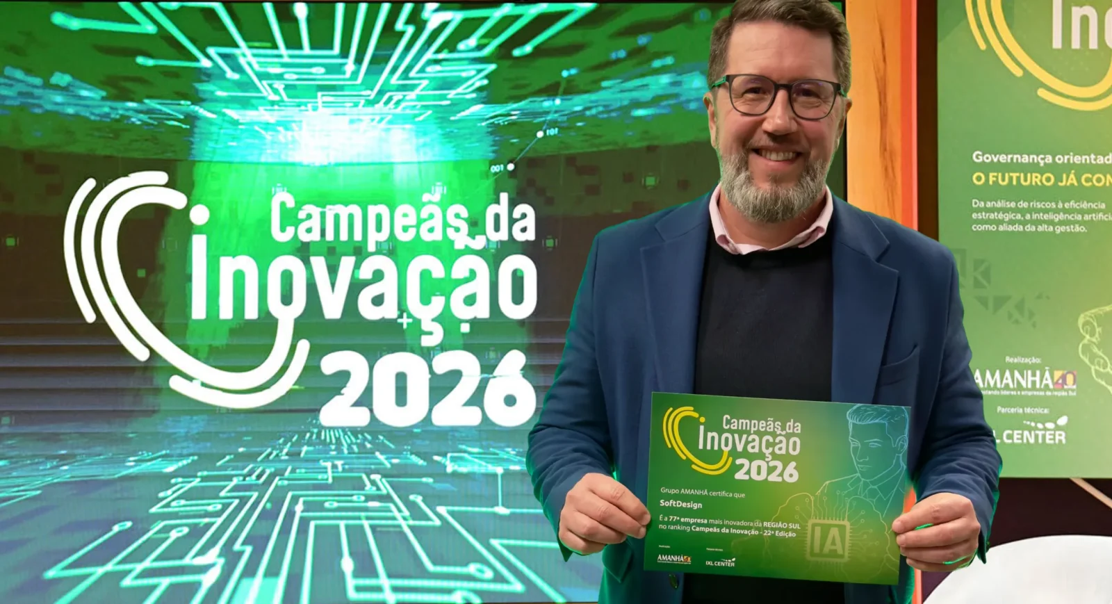 Imagem promocional com um homem sorrindo ao lado de um banner com o texto “Campeões da Inovação 2026”, segurando um certificado na mesa de evento.